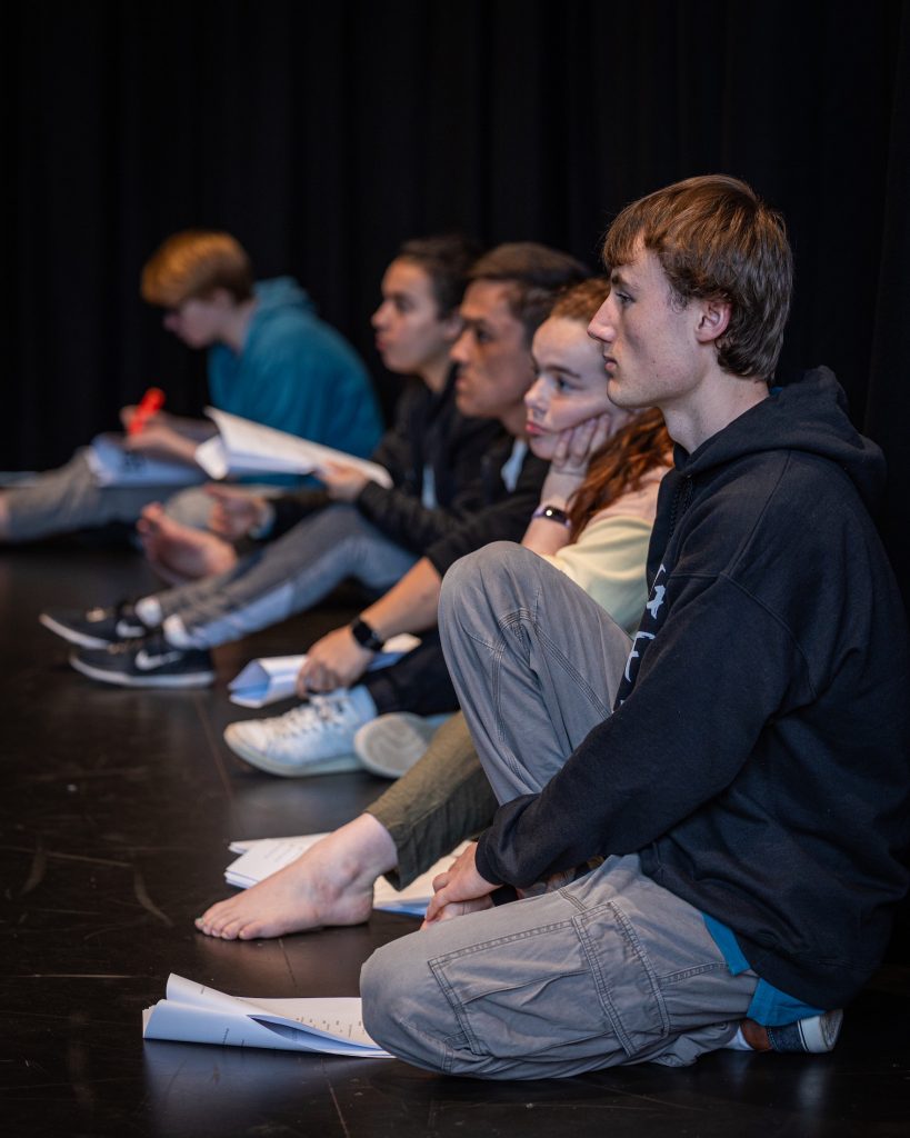 A group of young people sit on a stage floor, looking intently at the director (not pictured).